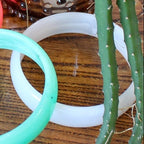 Two bangles, one green and one white, on a wooden surface with a cactus plant.